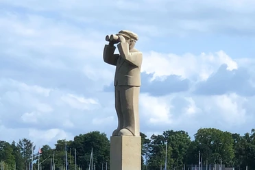 Ein Steindenkmal eines Hafenmeisters mit Fernglas, vor einem bewaldeten Hintergrund und Segelmasten.