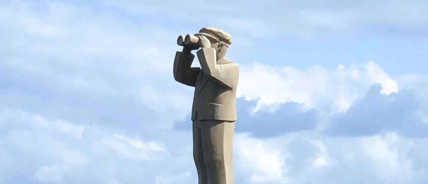 Ein Steindenkmal eines Hafenmeisters mit Fernglas, vor einem bewaldeten Hintergrund und Segelmasten.