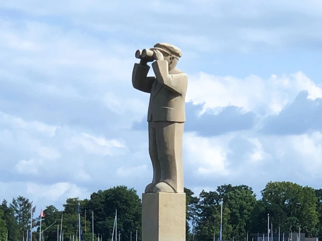 Der Hafenmeister Ein Steindenkmal eines Hafenmeisters mit Fernglas, vor einem bewaldeten Hintergrund und Segelmasten.