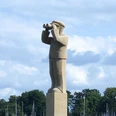 Der Hafenmeister Ein Steindenkmal eines Hafenmeisters mit Fernglas, vor einem bewaldeten Hintergrund und Segelmasten.