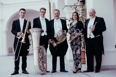 Fünf Musiker mit Blechblasinstrumenten in festlicher Garderobe stehen outdoor unter Arkaden.