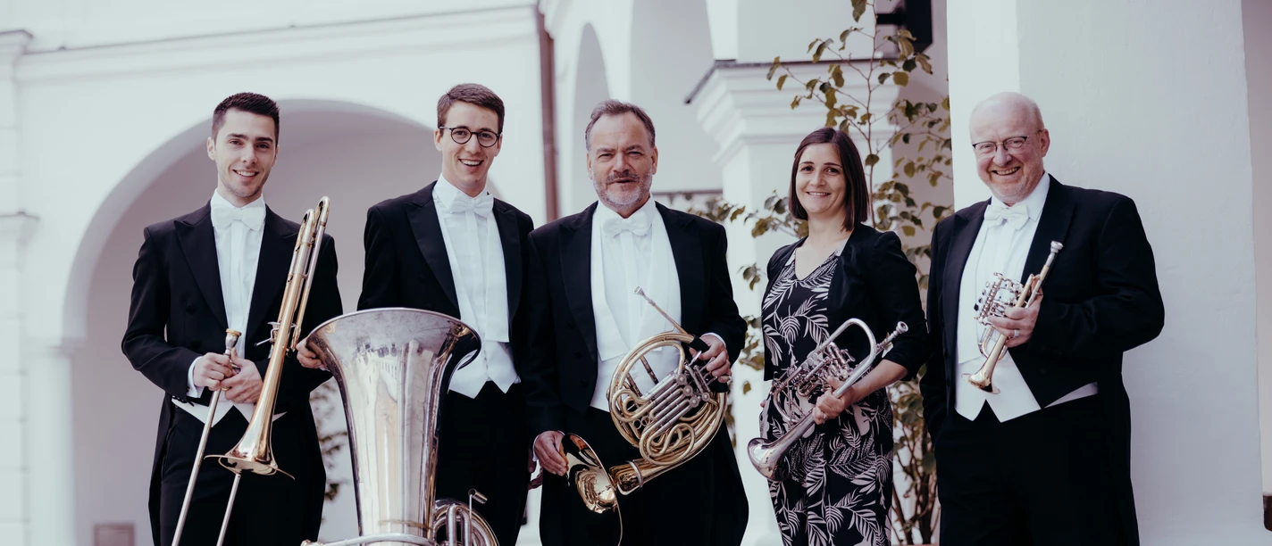 Fünf Musiker mit Blechblasinstrumenten in festlicher Garderobe stehen outdoor unter Arkaden.