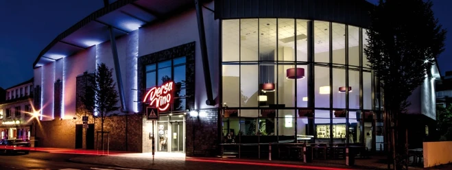 Außenansicht Modernes Kino bei Nacht, beeindruckende Glasfassade mit leuchtendem Kino-Logo und Bäumen davor.