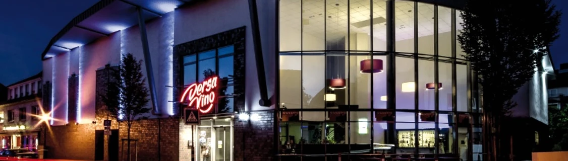 Außenansicht Modernes Kino bei Nacht, beeindruckende Glasfassade mit leuchtendem Kino-Logo und Bäumen davor.