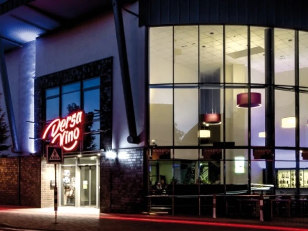 Außenansicht Modernes Kino bei Nacht, beeindruckende Glasfassade mit leuchtendem Kino-Logo und Bäumen davor.