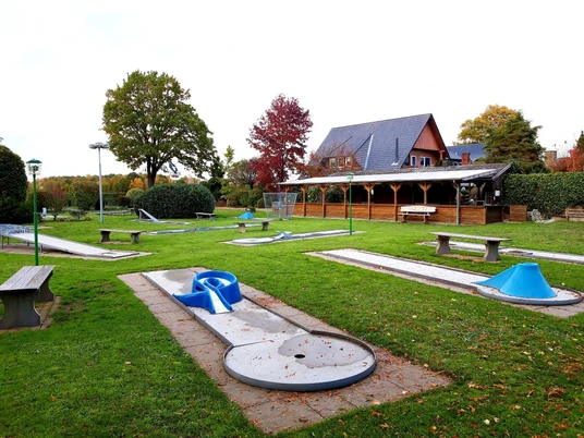 Minigolf Goldener Hecht Ein weitläufiger Minigolfplatz mit verschiedenen Bahnen, Bänken und einem hölzernen Pavillon im Hintergrund.