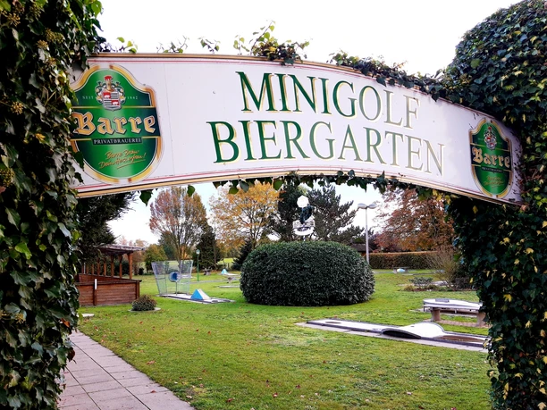 Minigolf Eingang zu einem Minigolf-Biergarten mit schattenspendenden Bäumen und einer gepflegten Anlage.