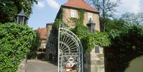 Schloss Petershagen Hohes, von Efeu bewachsenes Schlossgebäude mit rotem Ziegeldach und verziertem Eisentor im Vordergrund.