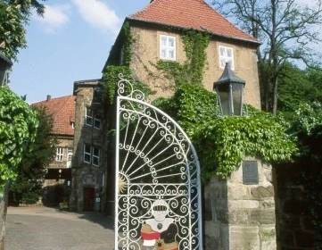 Schloss Petershagen Hohes, von Efeu bewachsenes Schlossgebäude mit rotem Ziegeldach und verziertem Eisentor im Vordergrund.