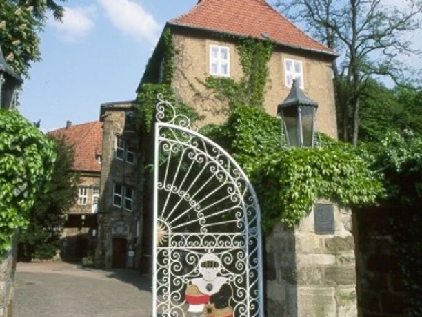 Schloss Petershagen Hohes, von Efeu bewachsenes Schlossgebäude mit rotem Ziegeldach und verziertem Eisentor im Vordergrund.