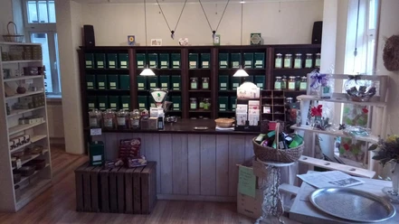 dat-teehus-tee-walsrode Das Bild zeigt das gemütliche Interieur von Dat Teehus in Walsrode. Ein rustikaler Verkaufstresen steht im Vordergrund, eingerahmt von Regalen voller Teedosen in warmen Holztönen. Dezente Beleuchtung und liebevoll arrangierte Dekorationen schaffen eine einladende Atmosphäre. Im Regal sind verschiedene Teesorten sichtbar, die zum Entdecken und Genießen einladen. Der Raum strahlt Wärme und Behaglichkeit aus, ideal für Tee-Liebhaber auf der Suche nach einem besonderen Geschmackserlebnis.