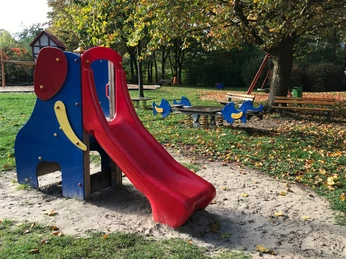 Bunter Spielplatz mit Rutsche und Wippe, umgeben von Bäumen und herbstlichen Laubblättern.