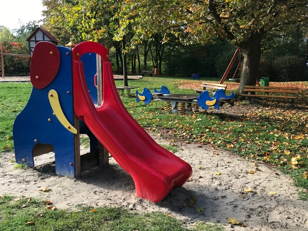 Die Elefantenrutsche und die Fischwippe Bunter Spielplatz mit Rutsche und Wippe, umgeben von Bäumen und herbstlichen Laubblättern.