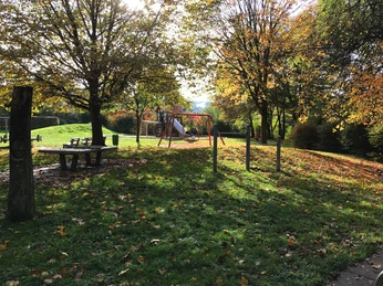 Spielplatz in herbstlicher Parklandschaft, umgeben von Laubbäumen und herabgefallenen Blättern.