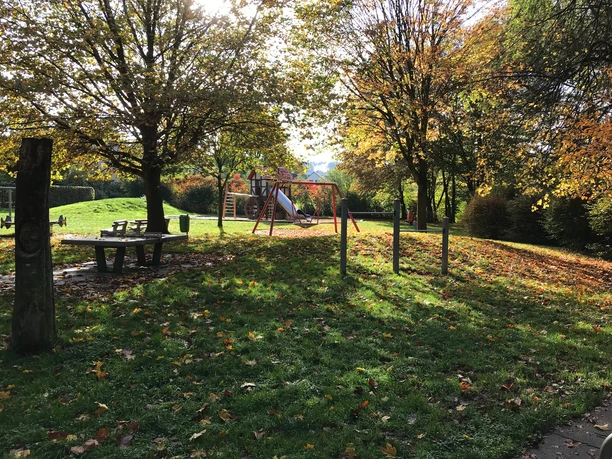 Spielplatz in herbstlicher Parklandschaft, umgeben von Laubbäumen und herabgefallenen Blättern.