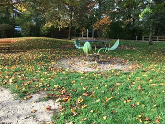 Ein grünes, schiffsförmiges Klettergerät auf einem Laubbedeckten Spielplatz an einem Herbsttag.