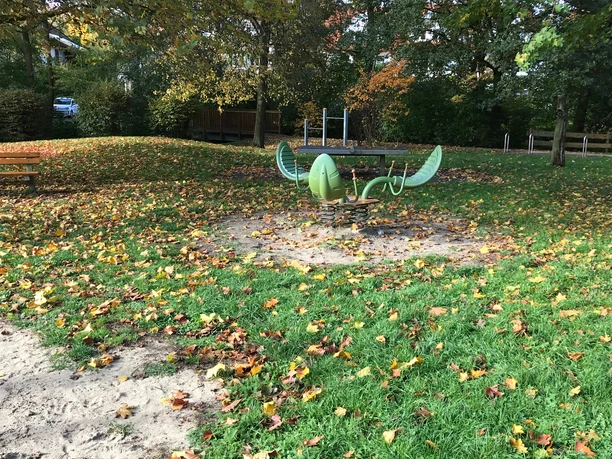 Ein grünes, schiffsförmiges Klettergerät auf einem Laubbedeckten Spielplatz an einem Herbsttag.