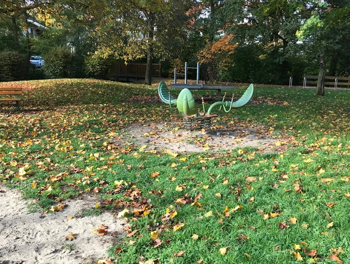 Ein grünes, schiffsförmiges Klettergerät auf einem Laubbedeckten Spielplatz an einem Herbsttag.
