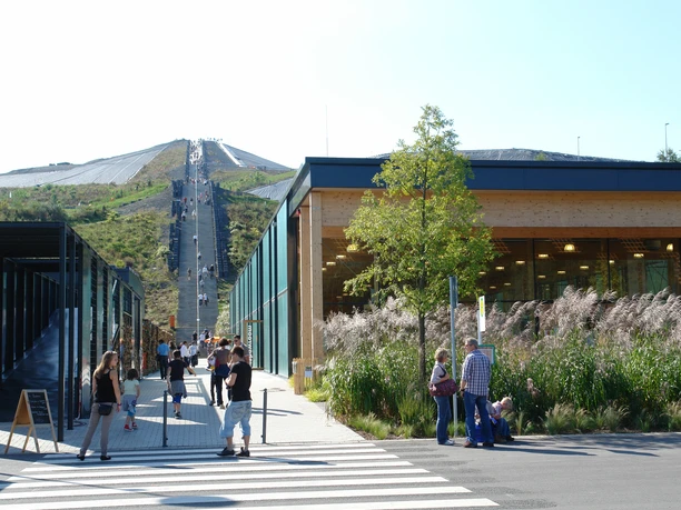 Blick auf den "schwarzen Kegel" von :metabolon Menschenmengen schlendern bei Sonnenschein entlang moderner Gebäude, im Hintergrund ein begrüntes Hügelplateau.