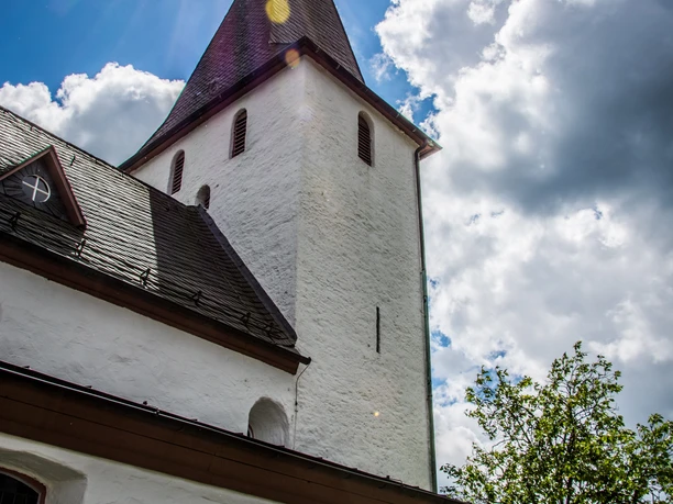 Bonte Kerke Lieberhausen Historische Kirche unter blauem Himmel mit Sonnenstrahlen durch Wolken, umgeben von Grün.