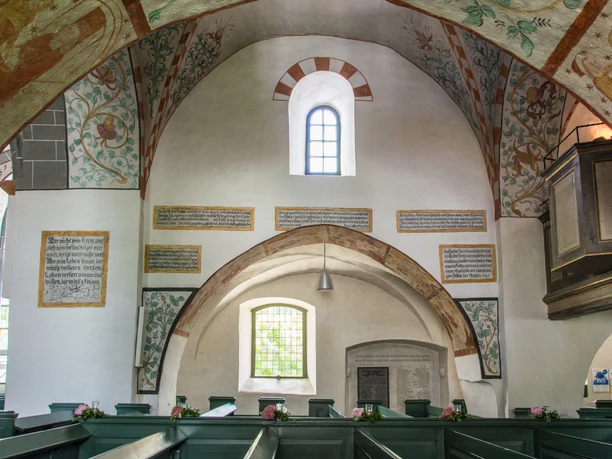 Innenansicht Innenansicht eines Kirchenraums mit bemalter Decke, weißen Wänden, Holzbänken und Fenstern.