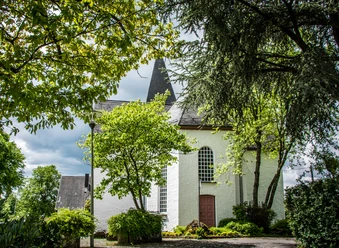 Bonte Kerke Lieberhausen Weiße Kirche in grüner Landschaft mit spitzem Turm, umrahmt von Bäumen; bewölkter Himmel.