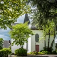 Bonte Kerke Lieberhausen Weiße Kirche in grüner Landschaft mit spitzem Turm, umrahmt von Bäumen; bewölkter Himmel.