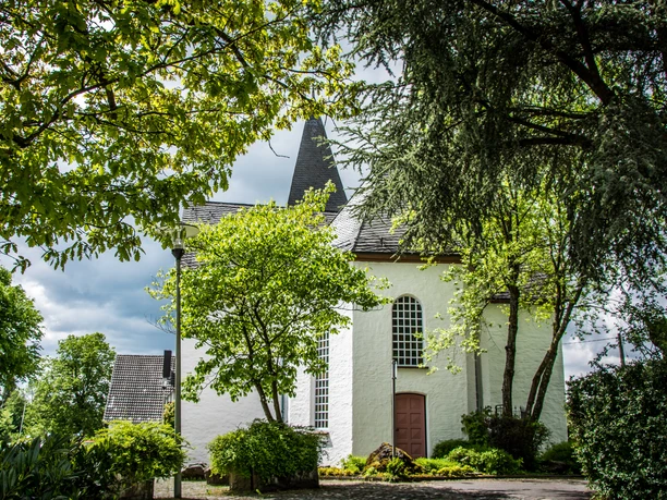 Bonte Kerke Lieberhausen Weiße Kirche in grüner Landschaft mit spitzem Turm, umrahmt von Bäumen; bewölkter Himmel.