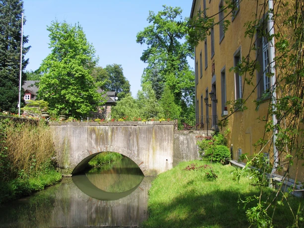 Schloss Heiligenhoven Historische Steinbrücke über einen kleinen Wassergraben, umgeben von grüner Natur und Gebäuden.