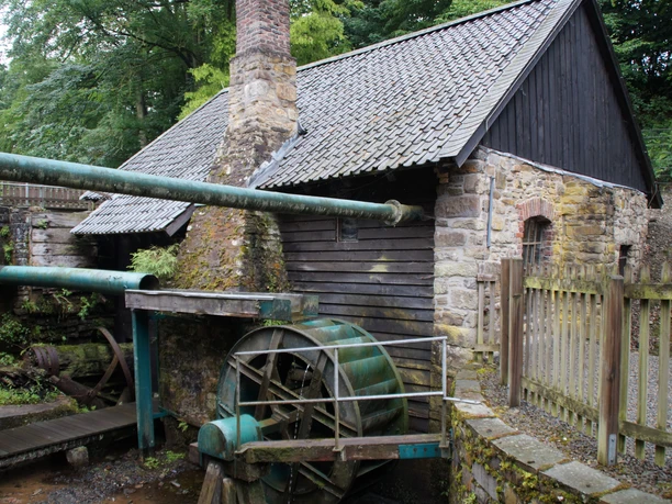 Wasserrad am Oelchenshammer Steinmühle mit Holzwasserrad und Moos am Gebäude, umgeben von Bäumen und Holzzaun im Freien.