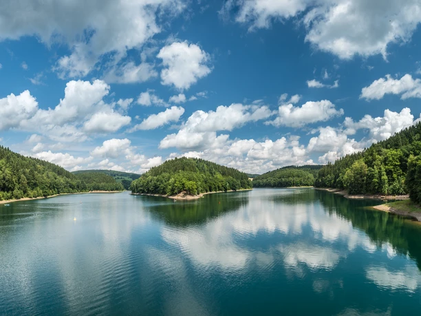 Blick auf die Aggerinsel Panorama eines Sees in Gummersbach, umgeben von bewaldeten Hügeln und reflektierenden Wolken im Wasser.