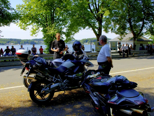 Motorradfahrer an der Bever Drei Motorradfahrer stehen gesellig bei ihren Maschinen vor einem See mit grünen Bäumen im Hintergrund.