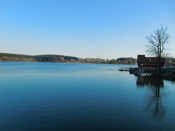 ein Gefühl wie in Skandinavien Klarer, ruhiger See mit spiegelndem Wasser, umgeben von Wald und einem Haus am Ufer unter blauem Himmel.