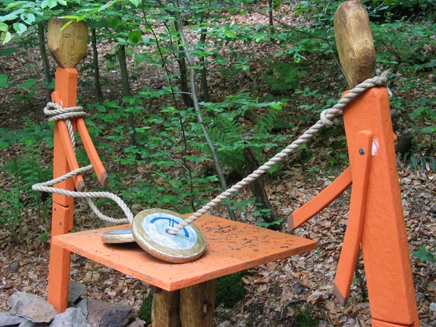 Mediendatei Der Waldparcours zeigt kunstvoll geschnitzte Figuren, die durch Seile verbunden sind, in einem Wald.