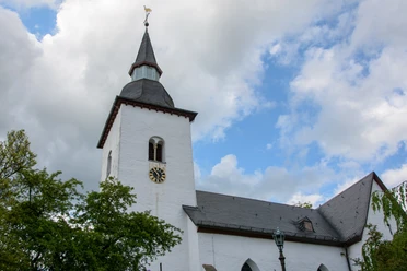 Bonte Kerke Marienberghausen Weiße Kirche mit spitzem Turm, Uhr und Hahn, von Bäumen umgeben, vor bewölktem Himmel.