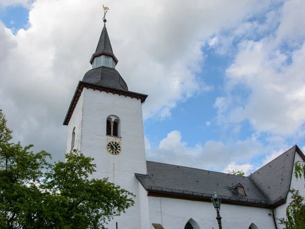Bonte Kerke Marienberghausen Weiße Kirche mit spitzem Turm, Uhr und Hahn, von Bäumen umgeben, vor bewölktem Himmel.