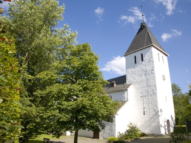 Bonte Kerke Müllenbach Weiße, historische Kirche mit spitzem Turm in grüner, baumreicher Umgebung unter blauem Himmel.