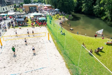 Aggerstrand Ründeroth Luftaufnahme eines lebhaften Strandvolleyballspiels neben einem Fluss, mit vielen Menschen und Zelten.
