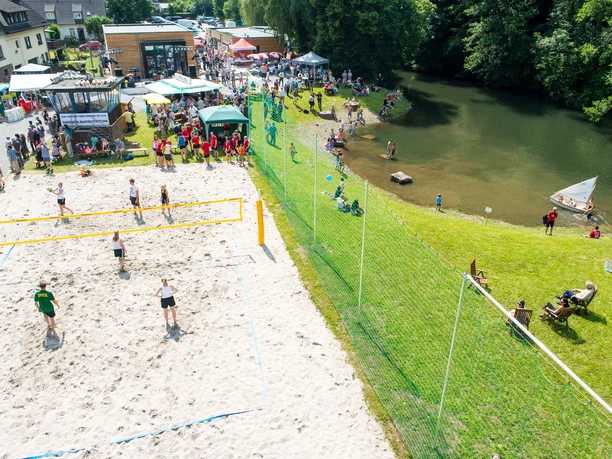 Aggerstrand Ründeroth Luftaufnahme eines lebhaften Strandvolleyballspiels neben einem Fluss, mit vielen Menschen und Zelten.