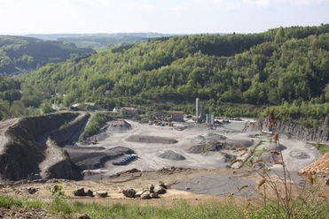 Blick in den Grauwacke-Steinbruch Steinbruch in grüner Hügellandschaft, mit Schotterfeldern und Industrieanlagen im Tal gelegen.