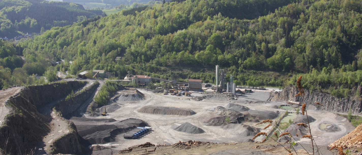 Blick in den Grauwacke-Steinbruch Steinbruch in grüner Hügellandschaft, mit Schotterfeldern und Industrieanlagen im Tal gelegen.