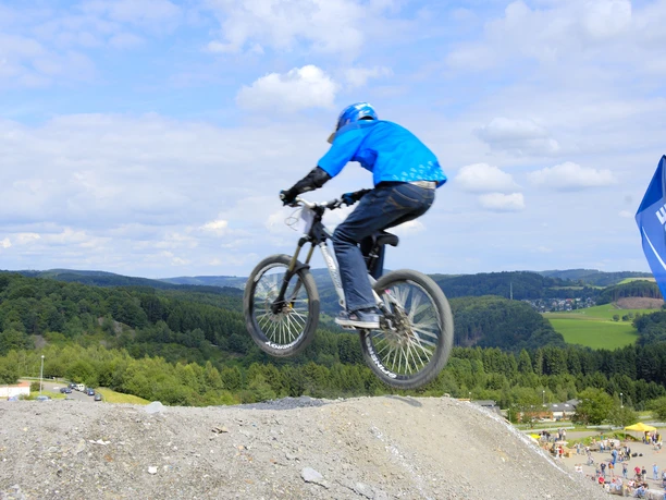 Radevent auf :metabolon Mountainbiker in blauem Outfit springt über Erdanhöhe vor bewaldeter Landschaft und blauem Himmel.