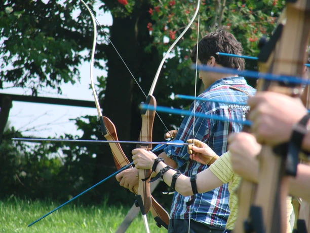 Mediendatei Mehrere Personen beim Bogenschießen im Freien, grüne Wiese und Bäume im Hintergrund.