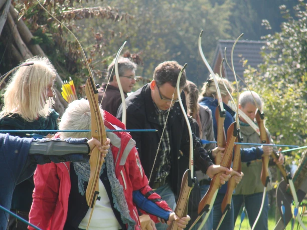 Arche e.V. Eine Gruppe von Menschen zieht Bögen bei einem Bogenschießkurs im Freien, umgeben von Natur.