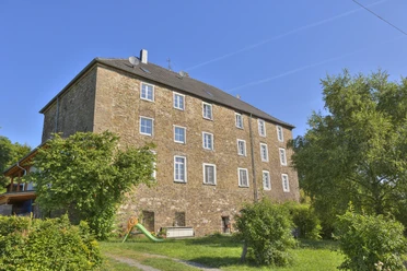 Burg Breidenbach Mehrstöckiges historisches Gebäude aus Naturstein, umgeben von Bäumen und einem kleinen Spielplatz.