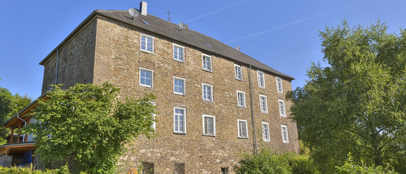 Burg Breidenbach Mehrstöckiges historisches Gebäude aus Naturstein, umgeben von Bäumen und einem kleinen Spielplatz.