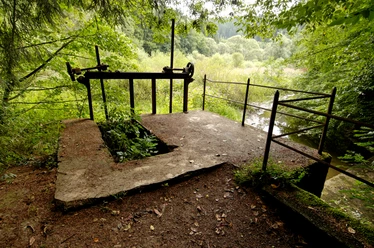 Burscheider Wehr Eine verlassene betonierte Plattform im Wald, umgeben von dichten Bäumen und einem dahinter liegenden Fluss.
