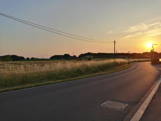 Domblick Breite Ländliche Straße bei Sonnenuntergang, umgeben von Feldern und Strommasten, sanfte abendliche Atmosphäre.