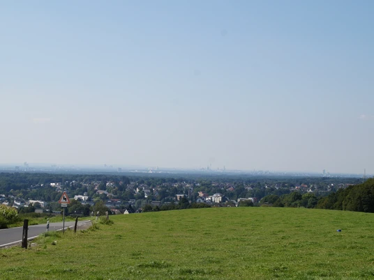 Domblick Schönrath Weitläufige grüne Wiese mit Blick über eine Stadtlandschaft, blauem Himmel und einigen Bäumen am Rand.