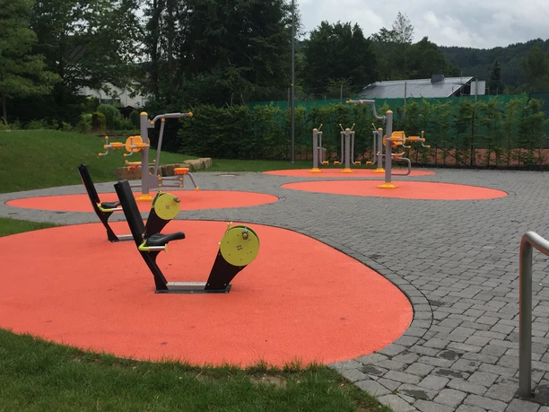 Freizeitanlage Bielstein Ein Fitnesspark mit Outdoor-Geräten auf orangefarbenen Flächen und grauem Pflaster vor einem Wald.